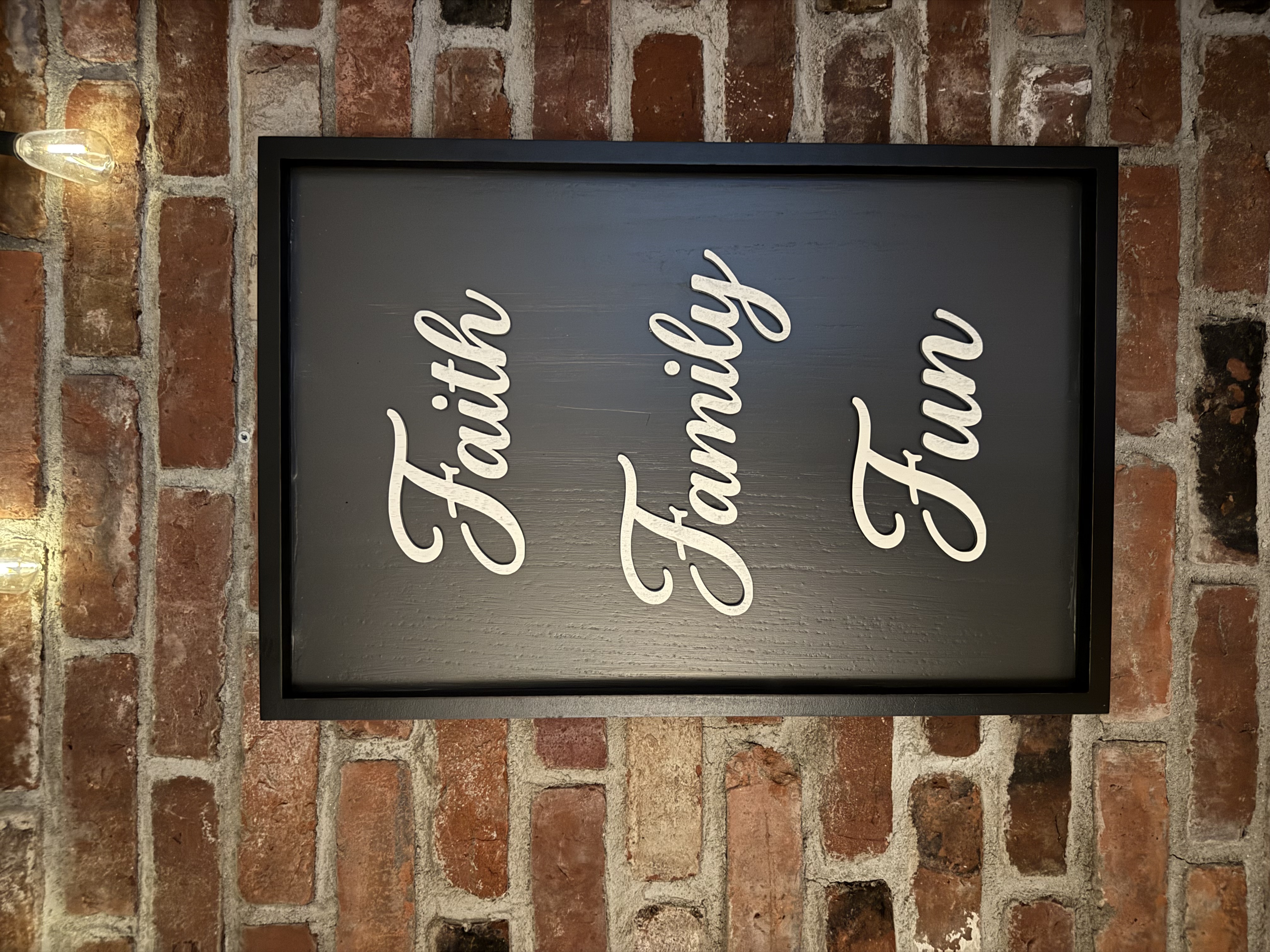 Framed black wood sign with Faith Family Fun in cream script on a brick wall